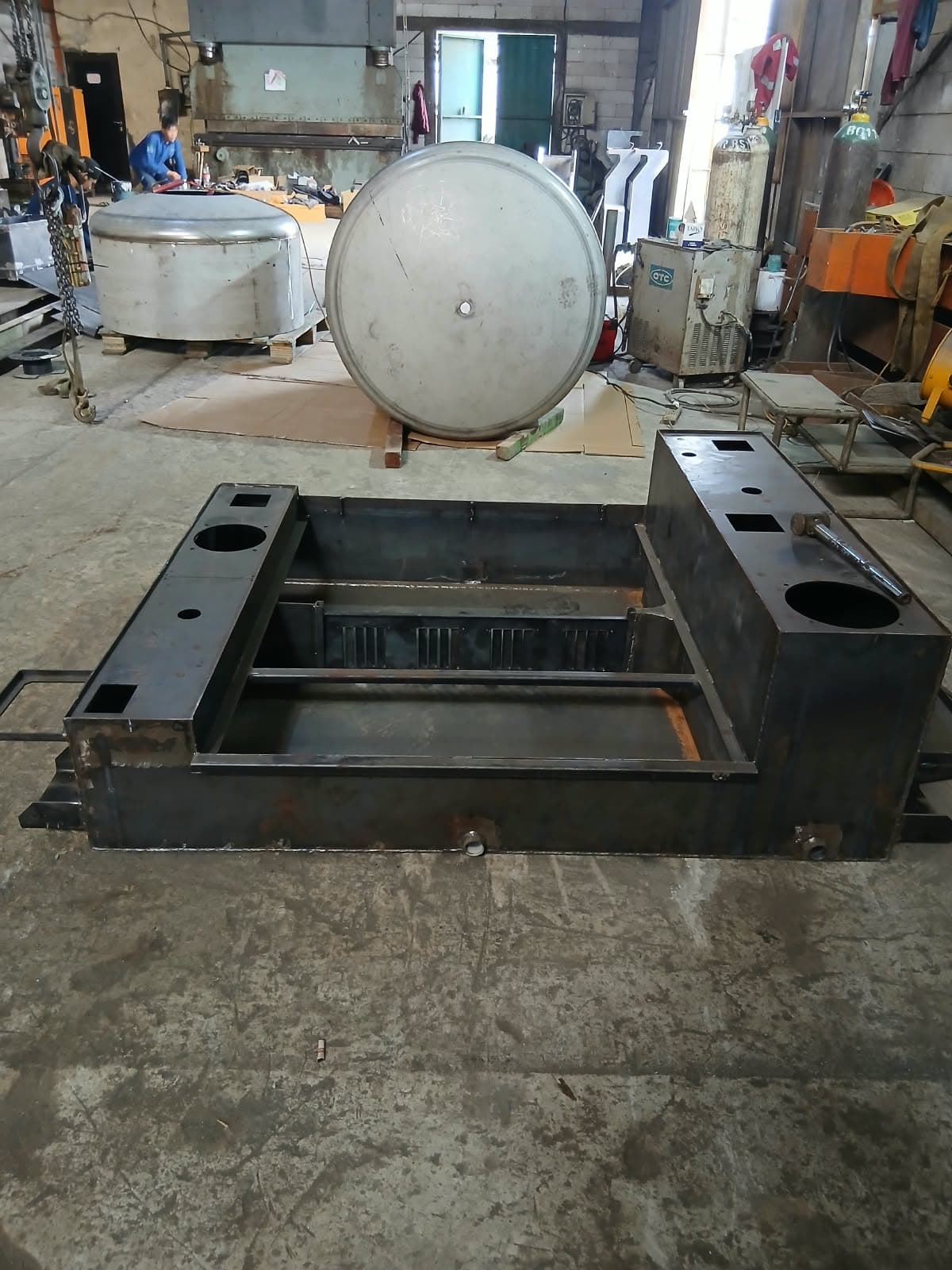 Rangka dasar baja hasil machining di lantai bengkel, dengan tutup tangki stainless di belakangnya